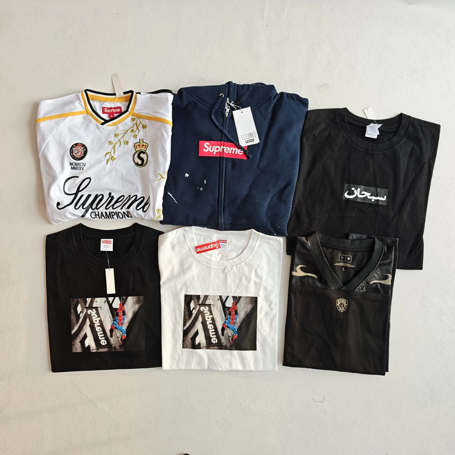 Flat-lay overview of a white-and-gold jersey, navy hoodie, dark logo tee, two graphic tees and a dark long-sleeve jersey.